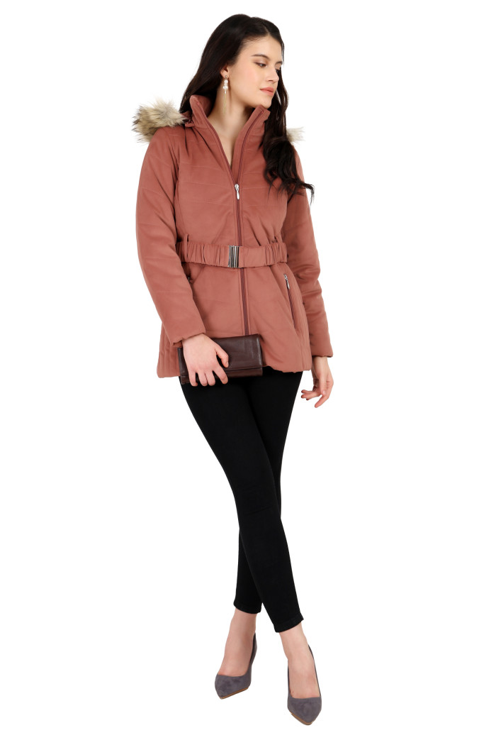 A woman stands confidently holding a clutch, styled in Coatsnmore’s onion pink velvet quilted jacket featuring a mandarin collar, removable hood, adjustable waistband zip closure, concealed zippered side pockets and black jeans and crossing her legs.